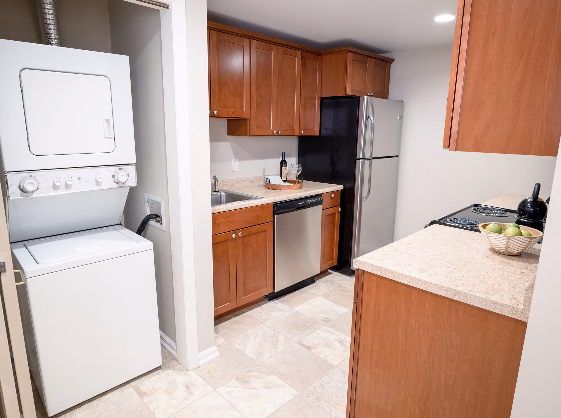 A compact kitchen area with wooden cabinets, a stainless steel refrigerator, dishwasher, and electric stove. To the left, there is a stacked washer and dryer unit in a small alcove. The countertops are light-colored, and there is a bowl of green apples and a black kettle on the counter.