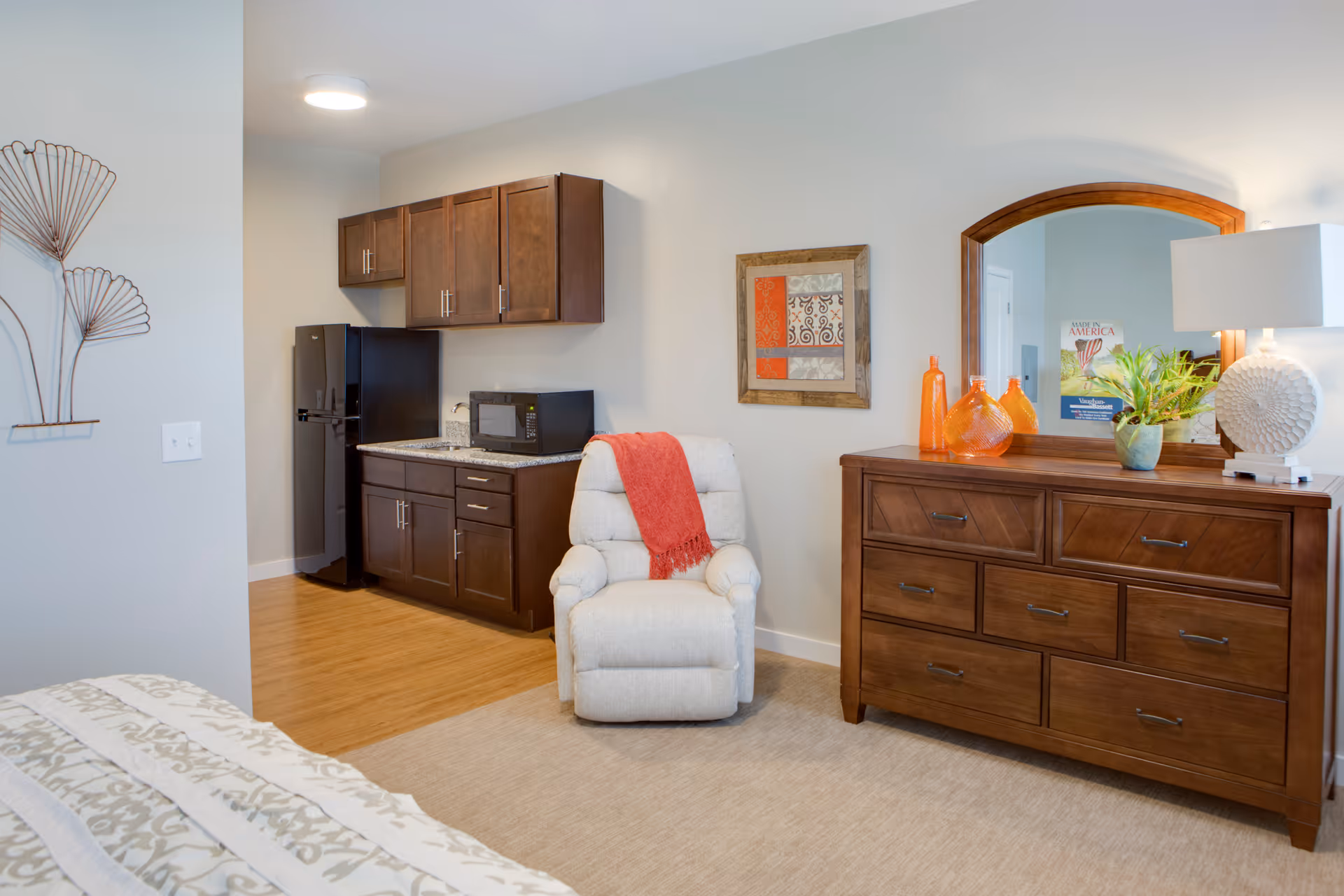 A cozy room featuring a small kitchenette with dark wood cabinets, a black refrigerator, and a microwave. In front of the kitchenette is a beige recliner chair with a red throw draped over it. To the right, there is a wooden dresser with a large mirror above it, decorated with orange glass bottles, a small plant, and a white lamp. Part of a bed with patterned bedding is visible in the foreground.