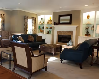 A cozy living room with two dark blue sofas, a beige armchair, a brown leather ottoman, and a fireplace with a mirror above it. Built-in shelves on either side of the fireplace display decorative items and are softly lit. The room has beige walls, carpeted floors, and windows with patterned curtains allowing natural light to enter.