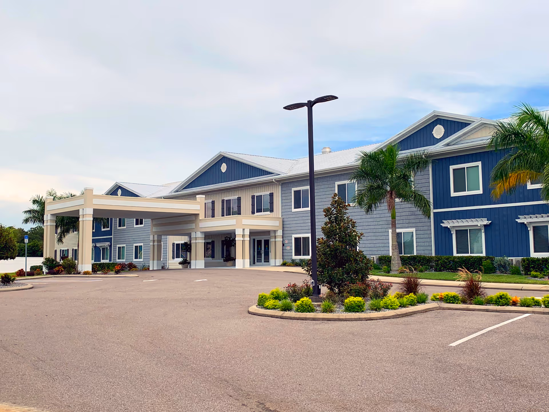 Exterior view of a two-story senior living facility building with blue and beige siding, a covered entrance, palm trees, landscaped greenery, and a parking lot in front under a partly cloudy sky.