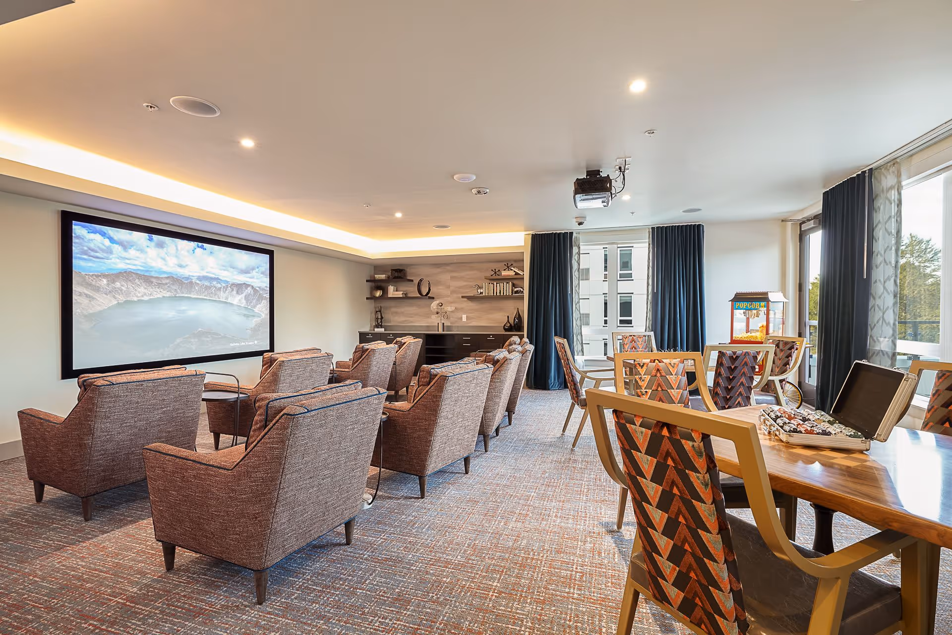 A bright community media room with rows of upholstered armchairs facing a large wall-mounted screen and tables with patterned chairs near windows.