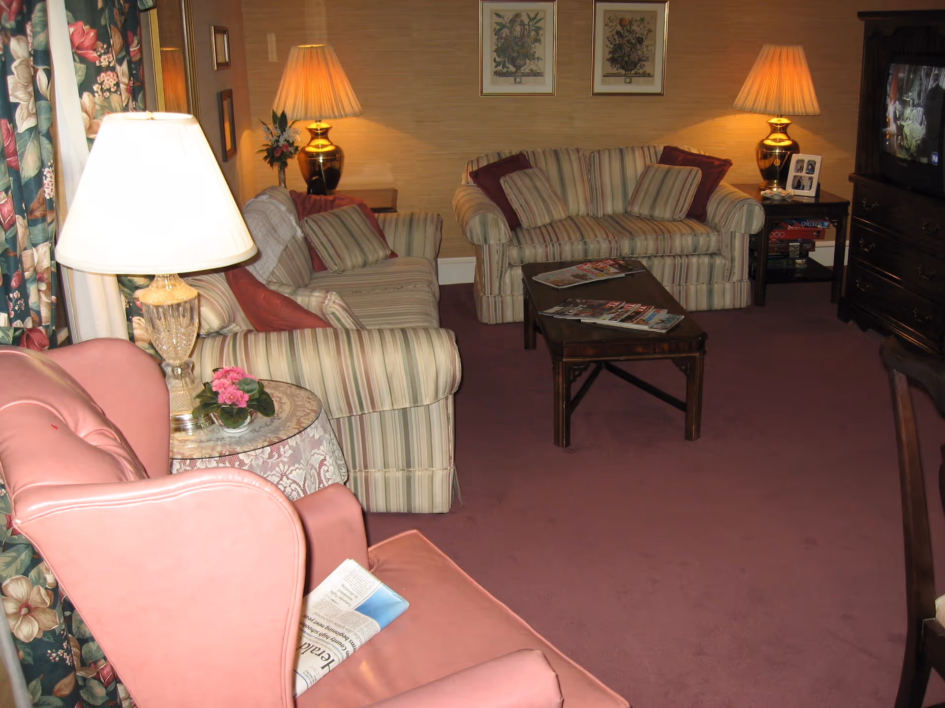 A cozy living room with two striped sofas, a pink armchair, a wooden coffee table with magazines, two table lamps with warm light, floral curtains, framed floral artwork on the wall, and a television on a wooden stand.