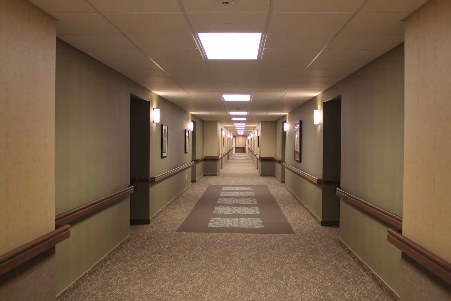 A long, well-lit hallway in a retirement community with beige walls, carpeted floor, handrails on both sides, and framed pictures hanging on the walls.