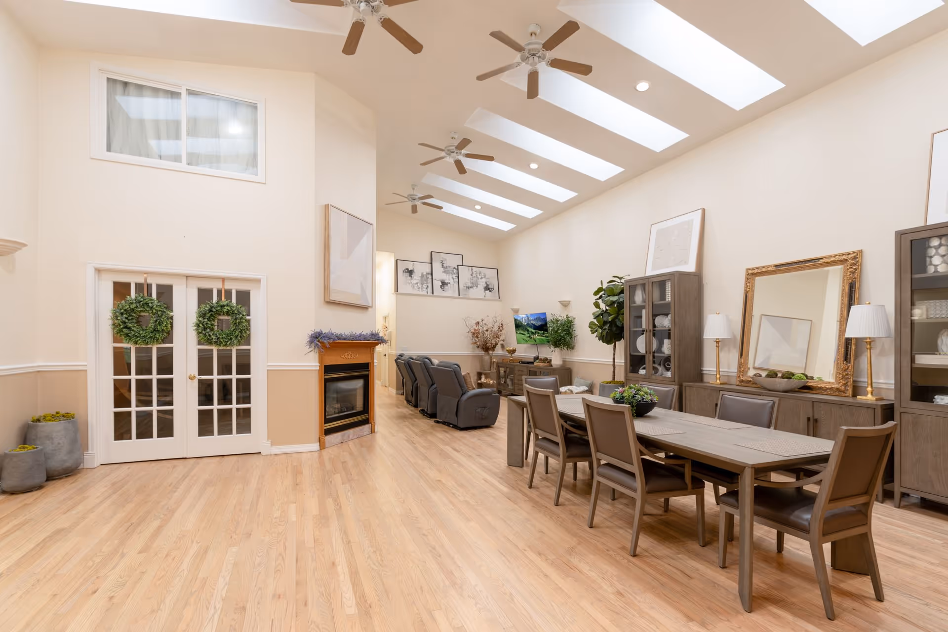 Bright communal dining and lounge area with a long dining table and chairs, skylights and ceiling fans, recliners by a fireplace, and decorative cabinets.