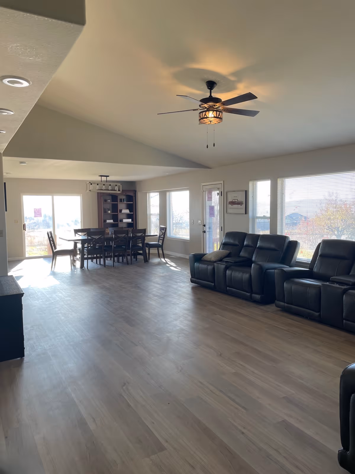 A spacious living and dining area with light wood flooring and large windows letting in natural light. The living area features black leather recliner chairs, and the dining area has a wooden table with chairs. A ceiling fan with a light fixture is mounted on the ceiling.