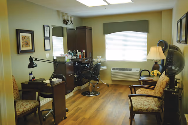 Interior view of a salon area in a senior living facility with a styling chair, hair dryer, wooden cabinets, two upholstered chairs, a table lamp, and framed artwork on the walls. The room has wood flooring and a large window with blinds.