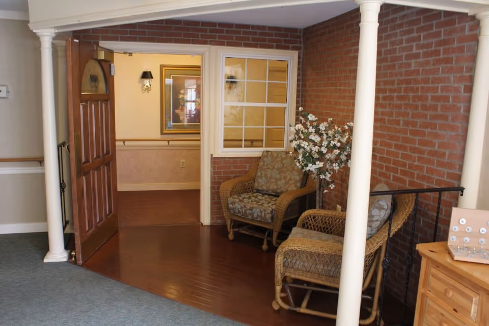 A cozy seating area inside a senior living facility with two wicker chairs featuring floral cushions, a small artificial flowering tree, and a wooden door open to a hallway with framed artwork and wall sconces. The area has a brick accent wall, wooden flooring, and white columns.