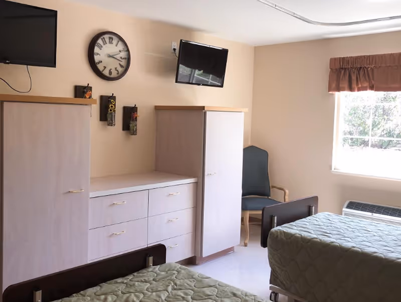 A nursing facility bedroom with two beds covered in green quilts, a wooden dresser with multiple drawers, two tall wardrobes, a wall clock, two wall-mounted televisions, a green cushioned chair, and a window with a brown valance letting in natural light.