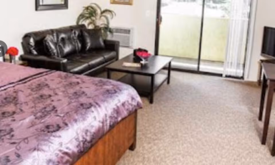 Studio-style bedroom with a bed in the foreground, a leather sofa and coffee table, and a sliding glass door to a balcony.