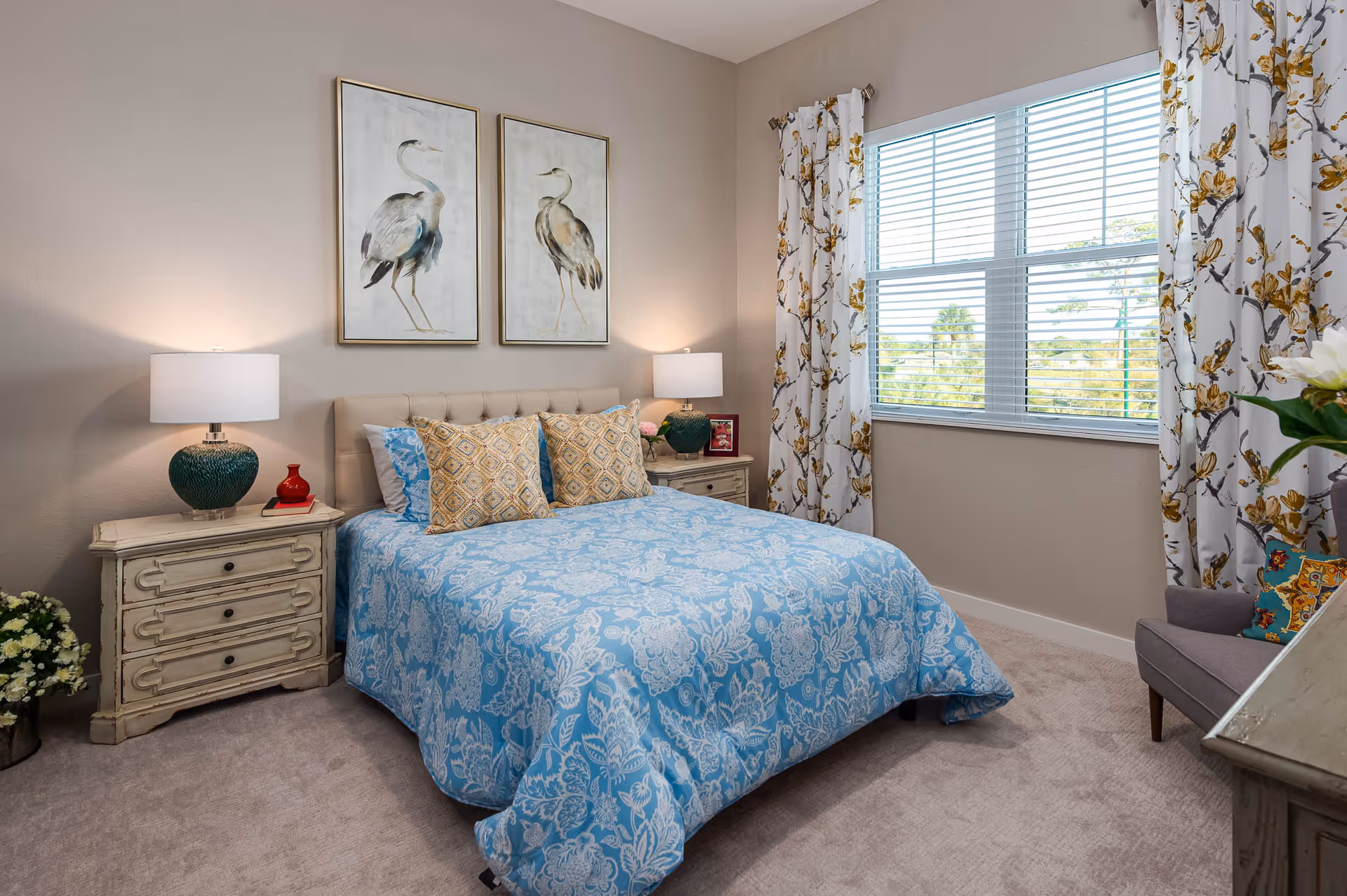 A cozy bedroom with a bed covered in a blue floral patterned bedspread and two decorative pillows. There are two matching nightstands on either side of the bed, each with a lamp. Above the bed, there are two framed pictures of birds. A window with floral curtains lets in natural light, and a gray armchair with a colorful pillow is placed near the window. The room has beige walls and carpeted flooring.