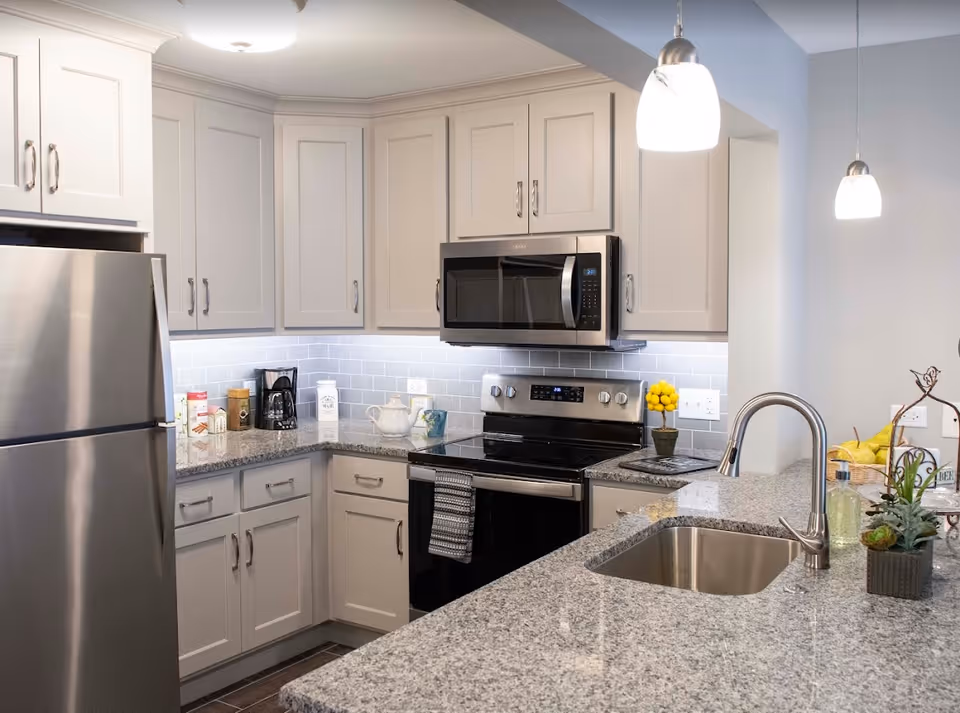 Modern kitchen with gray cabinets, stainless steel refrigerator, microwave, and stove. Granite countertops with a built-in sink and a few decorative items including a small potted plant and a bowl of lemons. Pendant lights hang above the counter.