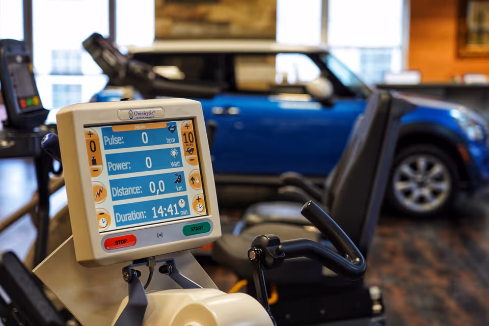 Close-up of a digital display on an exercise bike showing pulse, power, distance, and duration metrics in a fitness room with a blue car-shaped therapy device in the background.