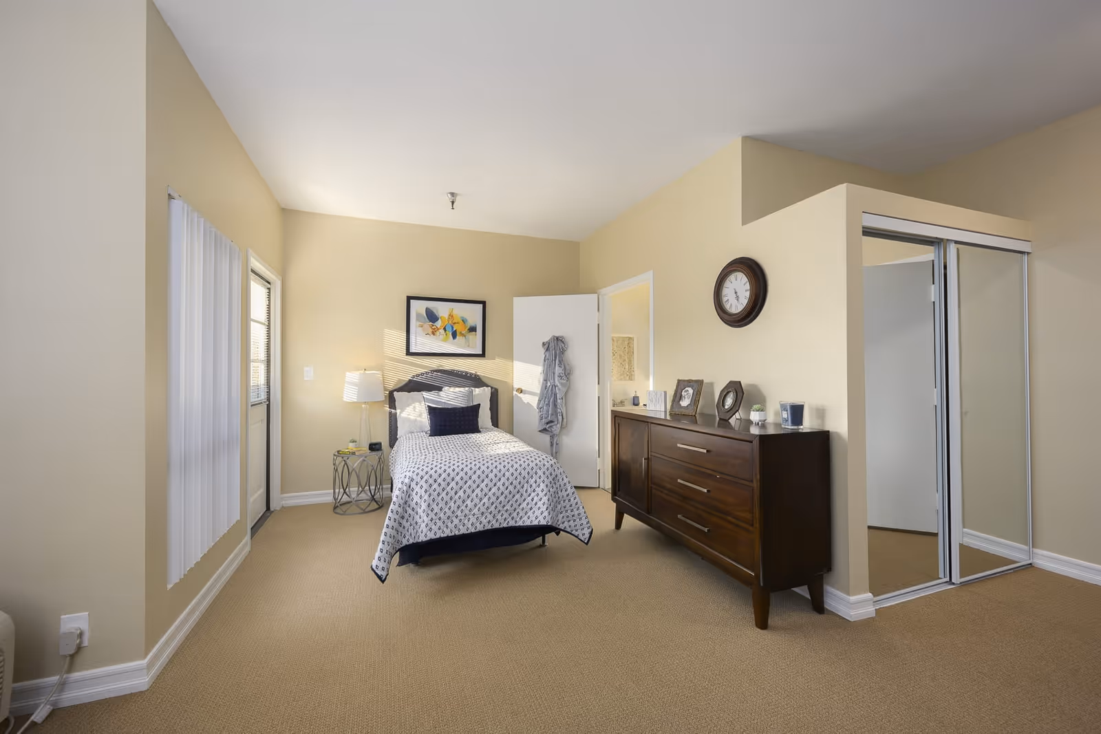 A neatly arranged bedroom with beige walls and carpet. The room features a single bed with a patterned white and black bedspread, a small round side table with a lamp, a wooden dresser with framed photos and a clock on the wall above it. There is a door leading to a bathroom and a mirrored sliding closet door.