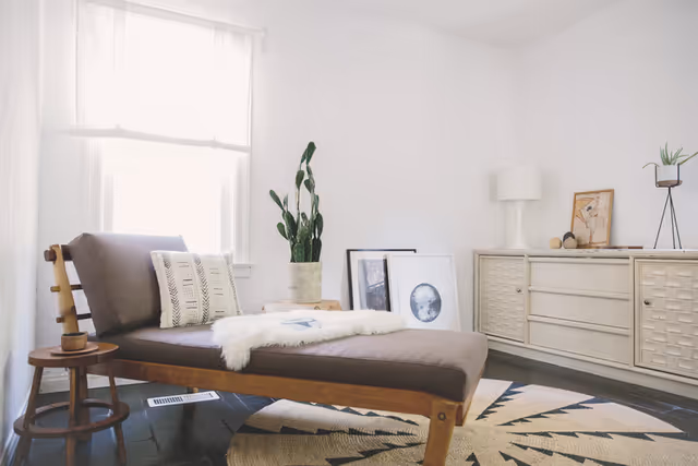A bright, minimalist living room with a wooden chaise lounge featuring a brown cushion and a white decorative pillow. A small round wooden side table with a cup sits next to the chaise. Behind it, there is a large window with sheer white curtains. On the floor is a round rug with a geometric pattern. Against the wall is a white sideboard with a lamp, framed pictures leaning against the wall, and a potted plant on top. The room has white walls and dark wooden flooring.
