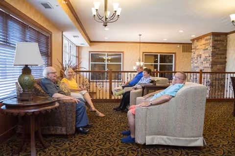 A cozy senior living common area with four elderly people sitting on comfortable armchairs and sofas, engaged in conversation. The room features warm lighting, large windows with wooden blinds, a decorative lamp on a side table, and a stone accent wall in the background.