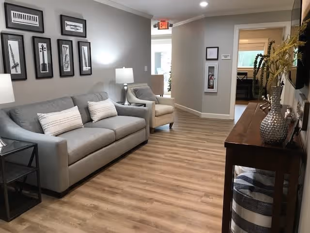 A cozy living room area in an assisted living facility featuring a gray sofa with two white pillows, a beige armchair, a side table with a lamp, and framed musical instrument artwork on the wall. The room has wood flooring and neutral-colored walls, with a wooden console table on the right holding decorative vases and a striped ottoman underneath. A hallway and an open door leading to another room are visible in the background.
