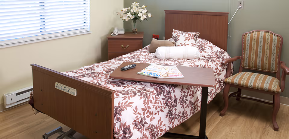 A single bed with a floral patterned bedspread and multiple pillows in a room with light green walls and wood flooring. Next to the bed is a wooden nightstand with a vase of white flowers and a telephone. A wooden chair with striped upholstery is positioned beside the bed. A small overbed table holds a TV remote and some magazines. A window with white blinds is on the left wall.