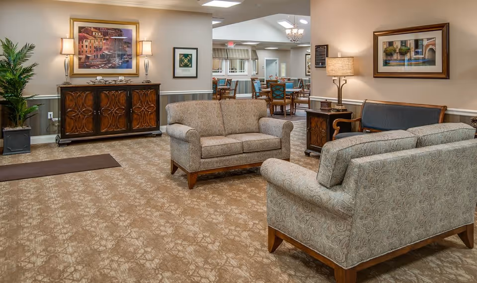 A cozy senior living facility common area with patterned carpet, two upholstered loveseats, a wooden bench with blue cushions, a wooden side table with a lamp, and a wooden cabinet with two lamps and framed artwork above it. In the background, there is a dining area with tables and chairs under a chandelier and skylight.