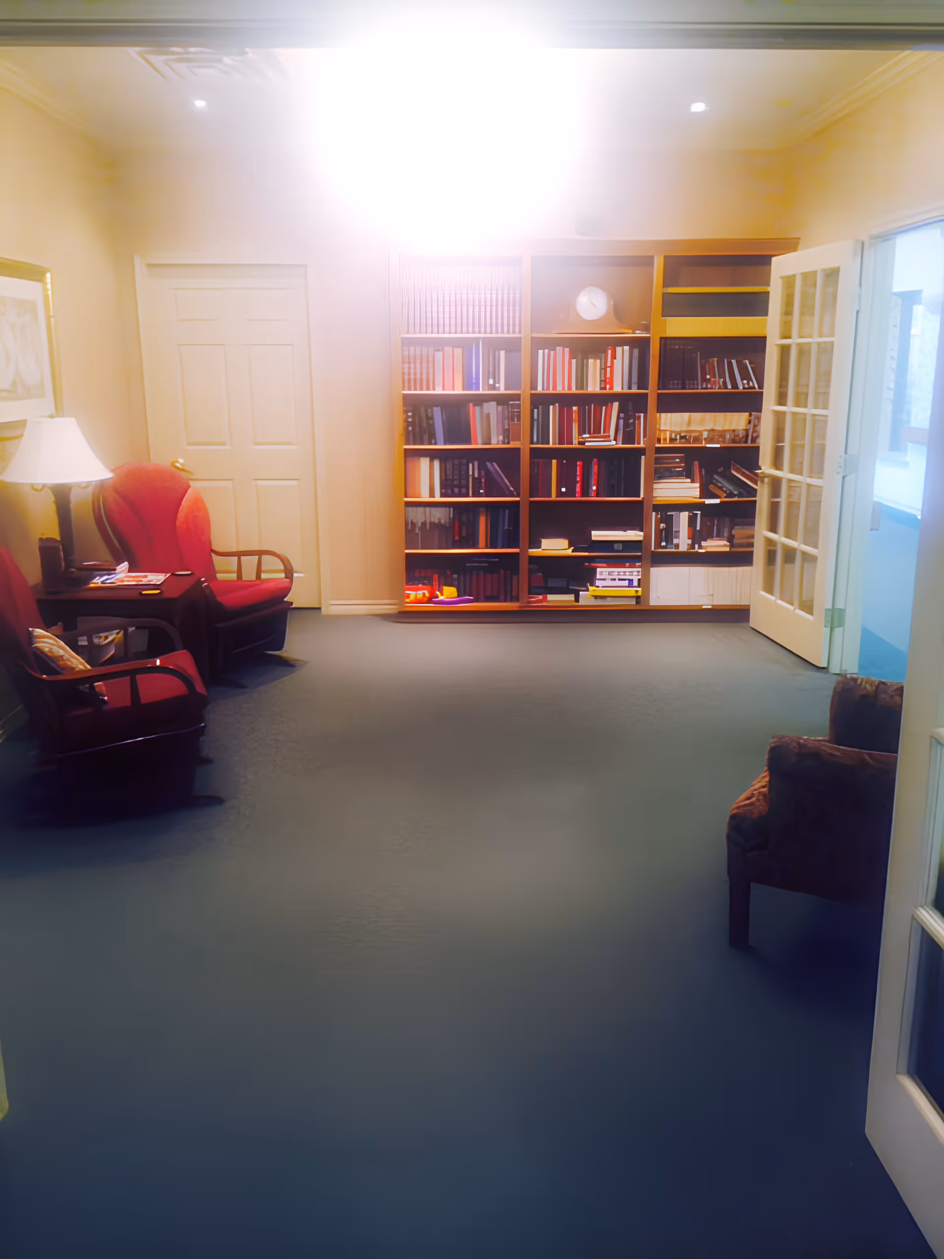 A cozy room with a large bookshelf filled with books against the back wall, two red armchairs with a small table and lamp between them on the left side, and a single armchair on the right near an open glass-paneled door. The room has carpeted flooring and soft lighting.