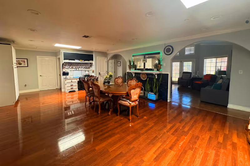Spacious interior of a senior living facility featuring a polished wooden floor, a dining area with a round wooden table and six ornate chairs, a fireplace with plants and decorative items, and an adjoining living room with comfortable seating and large windows allowing natural light.