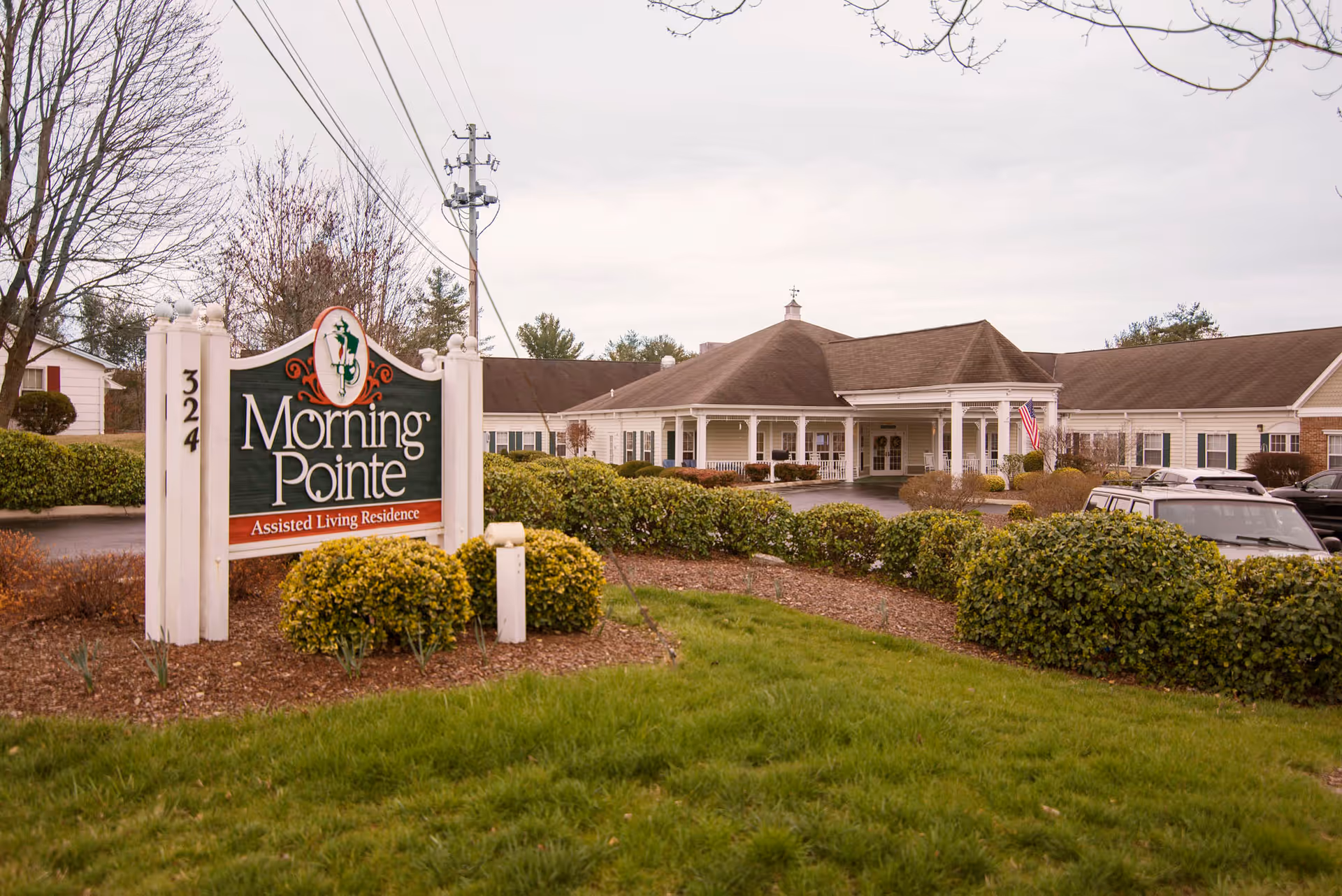 Exterior view of Morning Pointe Assisted Living Residence building with a large sign in the foreground displaying the facility name and address number 324. The building has a covered entrance, white columns, and an American flag near the entrance. There are bushes and green grass around the sign and building.