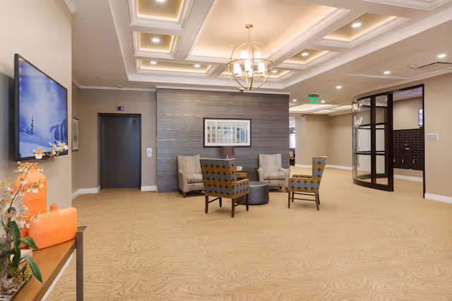 Bright, furnished lobby seating area with armchairs around a small table, a coffered ceiling and chandelier, wall-mounted TV, and an elevator door.