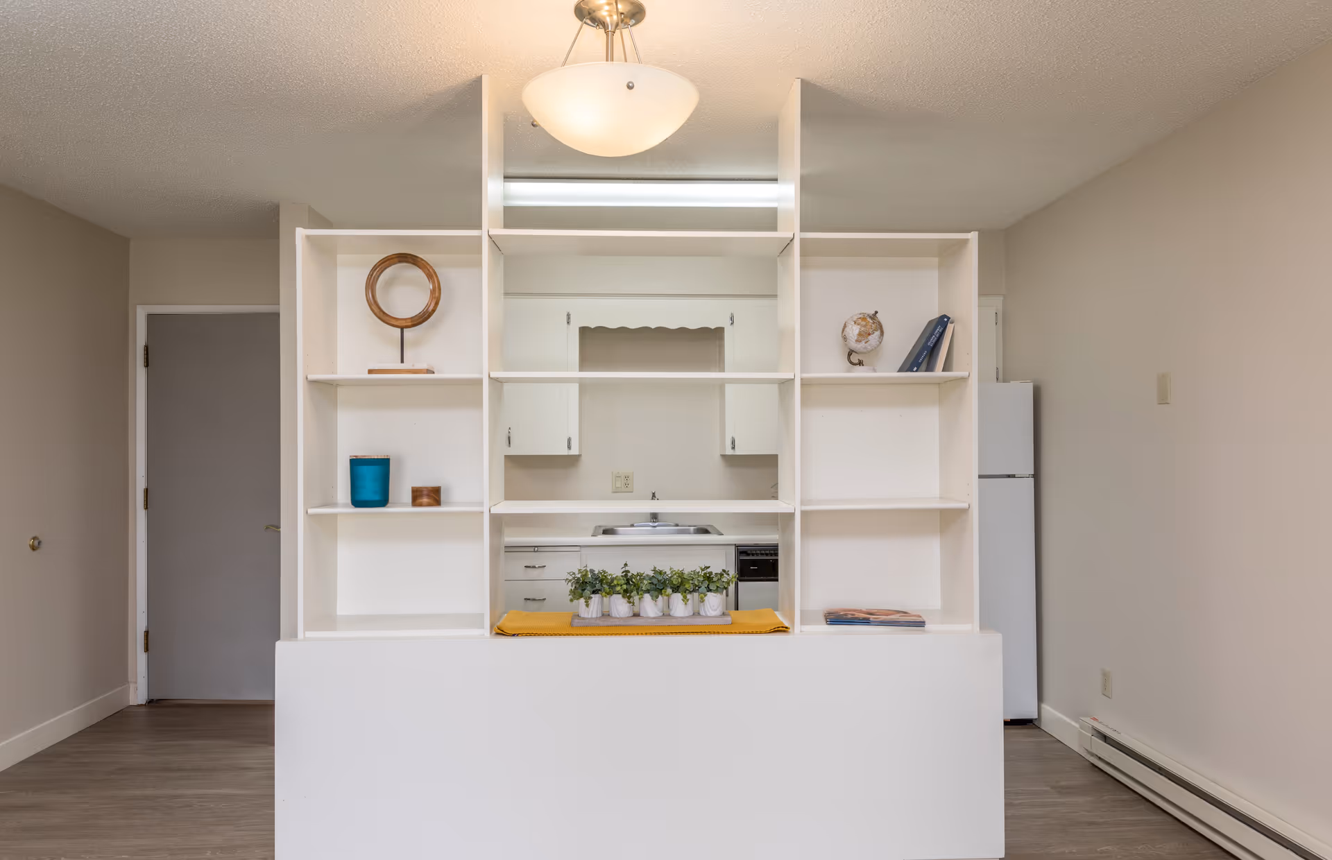 Interior view of a small kitchen area seen through a white open shelving unit with decorative items including a small globe, books, a round mirror, and potted plants. The kitchen has white cabinets, a sink, and a refrigerator on the right side. The walls are beige and the floor is wooden.