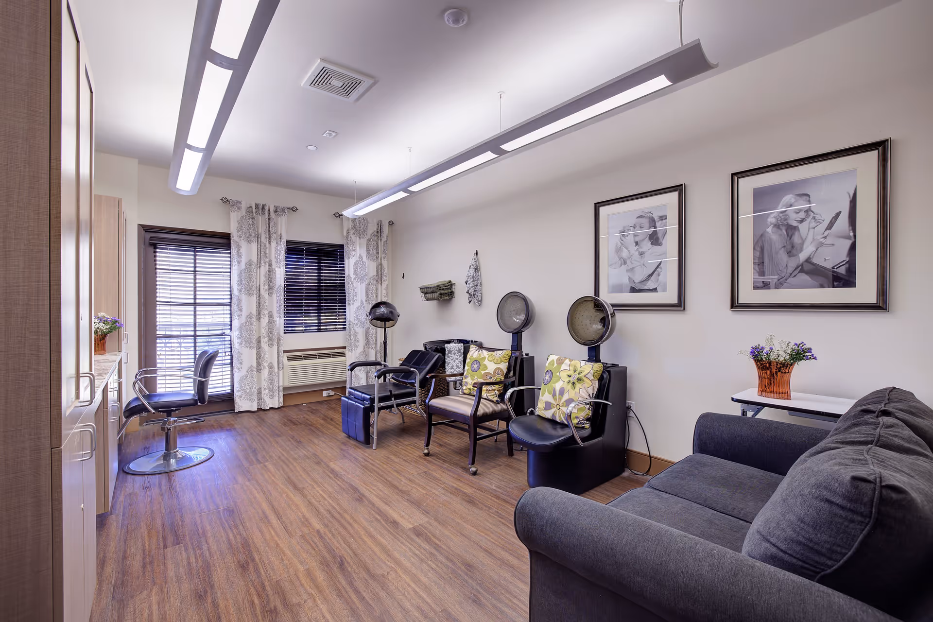 Interior of a senior living facility hair salon with wooden flooring, two hair drying chairs with hooded dryers, a black salon chair near a window with patterned curtains, a gray couch, and framed black and white photos on the wall.