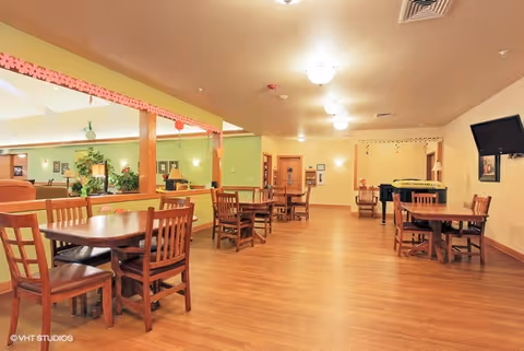 A spacious, well-lit common area with wooden floors and several wooden tables and chairs arranged throughout the room. The walls are painted in warm tones with some green accents, and there is a large mirror on one side reflecting part of the room. A flat-screen TV is mounted on the wall, and a piano is visible in the background. The ceiling has multiple light fixtures providing ample lighting.