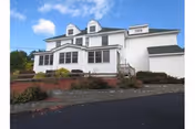 Exterior view of a large white multi-story house with green roof shingles, multiple windows, and a small porch with steps leading up to the entrance. The house is surrounded by landscaped bushes and a paved driveway under a blue sky with some clouds.