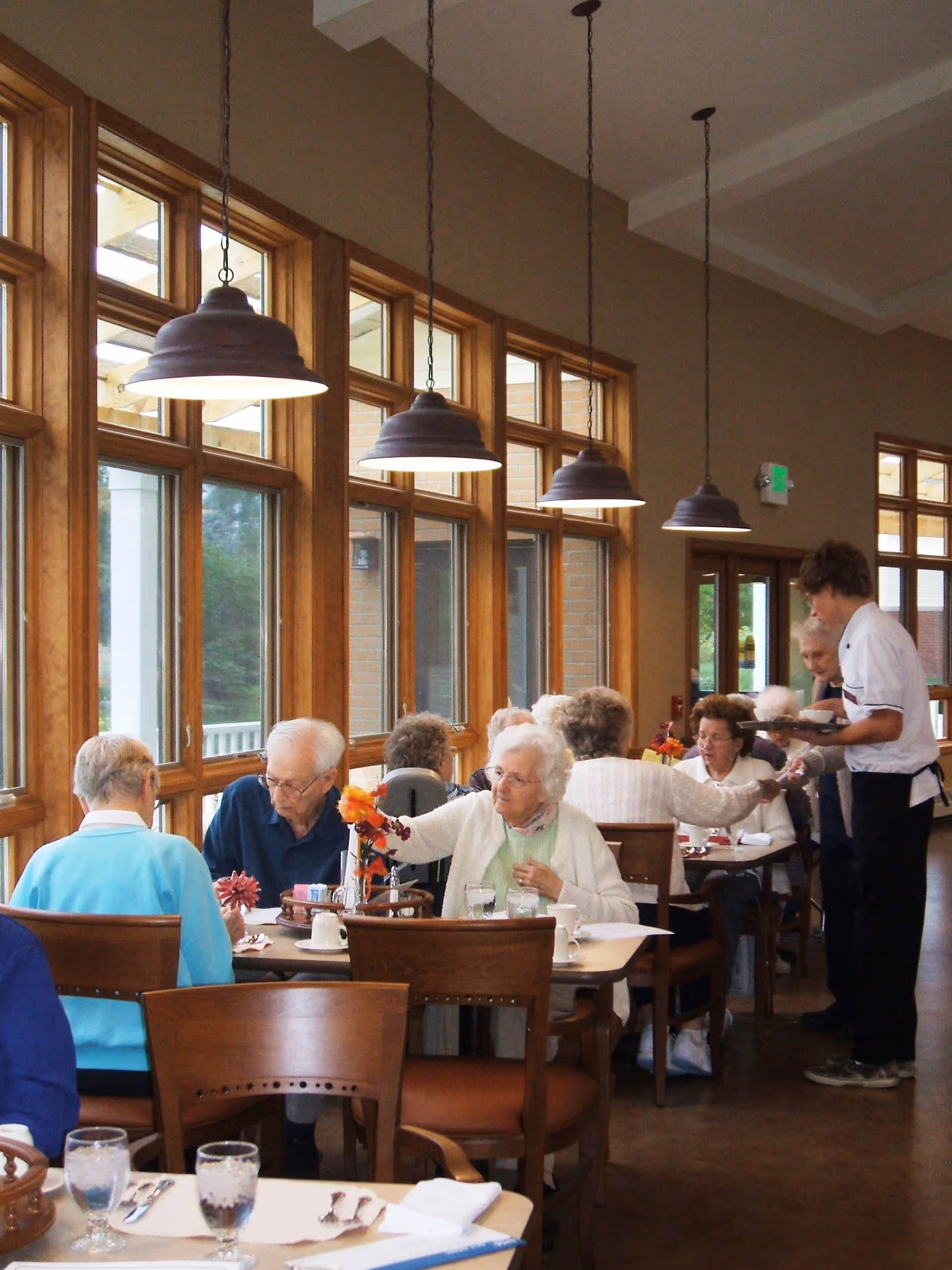 A group of elderly people sitting at tables in a dining room with large windows and hanging pendant lights. A server is attending to one of the tables, serving food and drinks. The room has wooden chairs and tables, and the atmosphere appears warm and social.