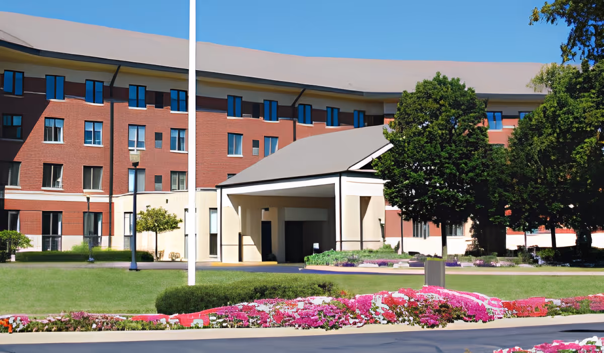 Exterior view of a multi-story brick building with a covered entrance, surrounded by green lawns, trees, and colorful flower beds under a clear blue sky.