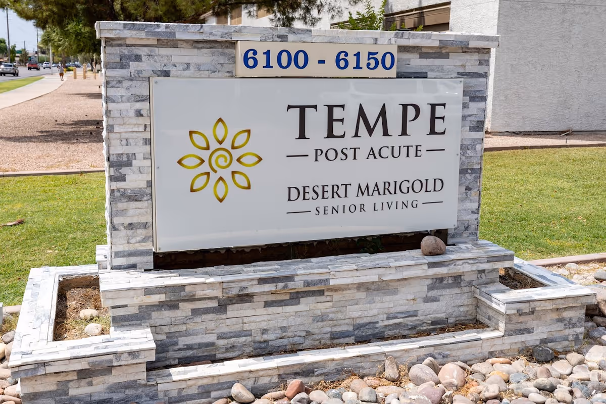 Stone sign for Tempe Post Acute Desert Marigold Senior Living with address numbers 6100 - 6150, set on a grassy area with rocks and a sidewalk nearby.