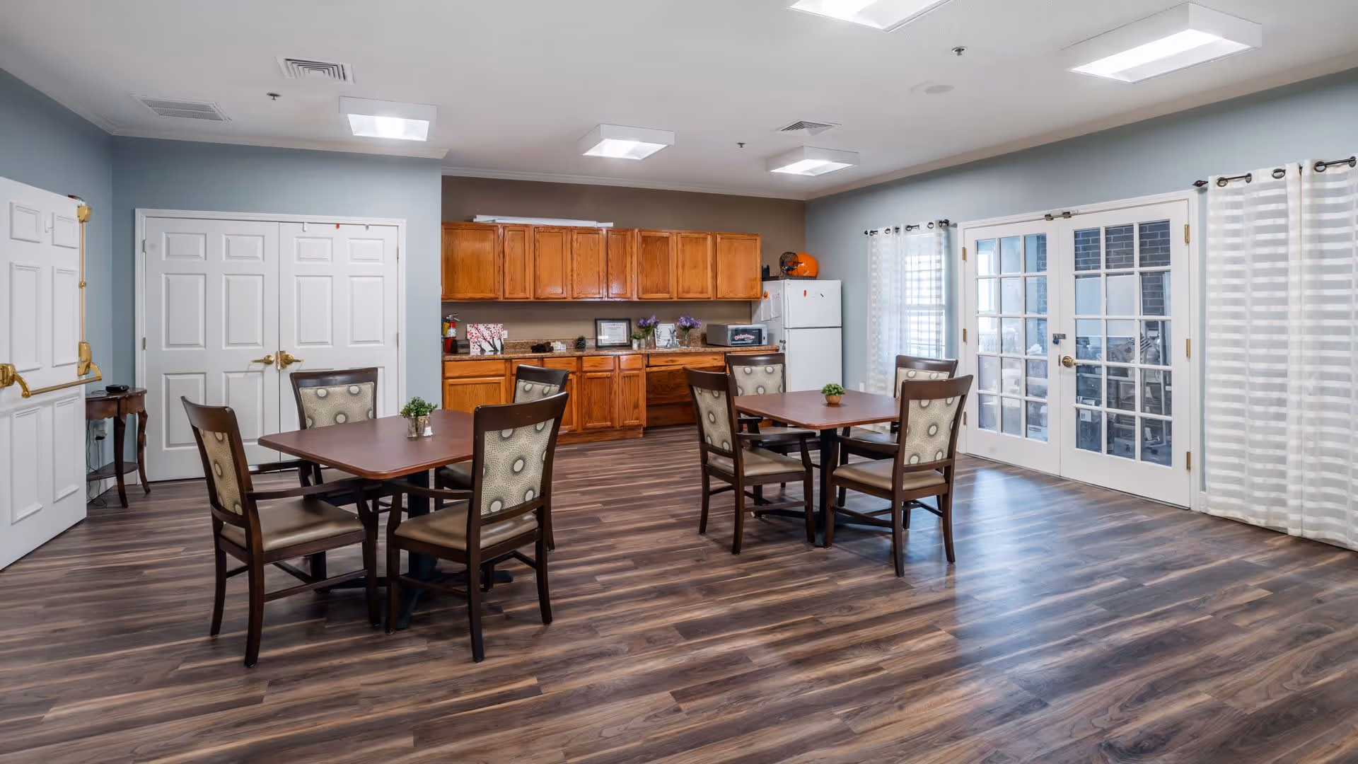 A bright and spacious dining room with two square wooden tables, each surrounded by four cushioned chairs. The room features wood flooring, light blue and brown walls, wooden cabinets, a white refrigerator, and large glass doors with sheer white curtains allowing natural light to enter.