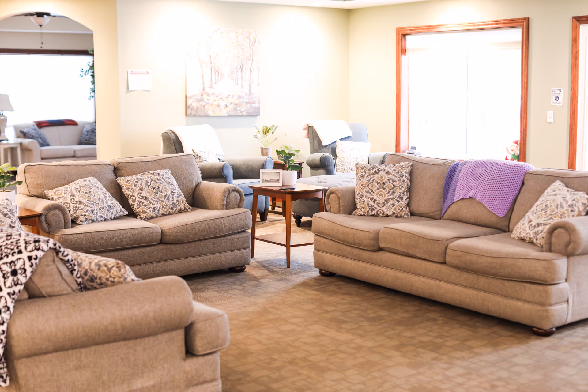 A cozy living room area in an assisted living facility with beige sofas and armchairs arranged around a wooden coffee table. The sofas have patterned cushions and a purple throw blanket. There is a large window with wooden trim letting in natural light, and a painting on the wall. The room has a warm and inviting atmosphere.