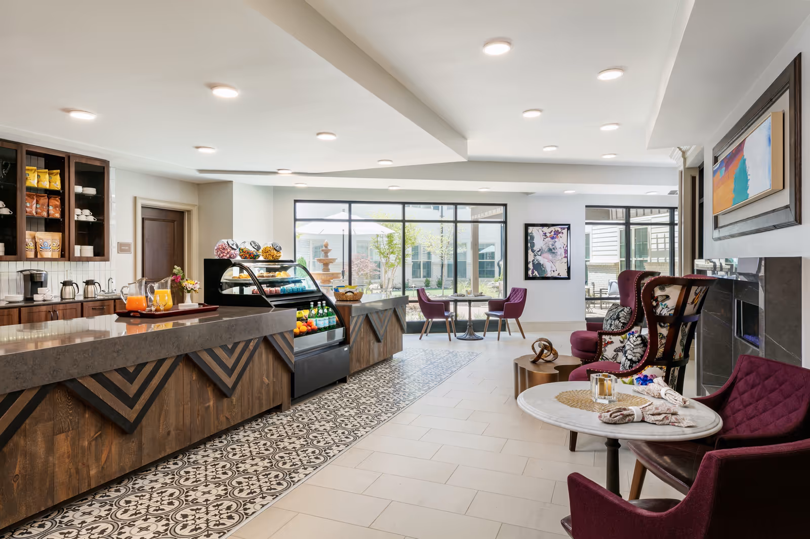 A bright and modern common area in a senior living facility featuring a long wooden counter with a display case of snacks and beverages. The floor has decorative tiles near the counter and light-colored tiles elsewhere. There are several purple upholstered chairs around small tables, a fireplace with artwork above it, and large windows showing an outdoor courtyard with a fountain and patio furniture.