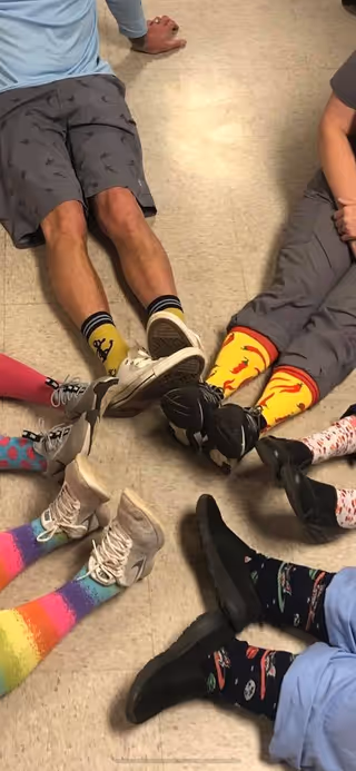 A group of people sitting on the floor in a circle with their legs extended inward, showing colorful and patterned socks and various types of footwear on a tiled floor.