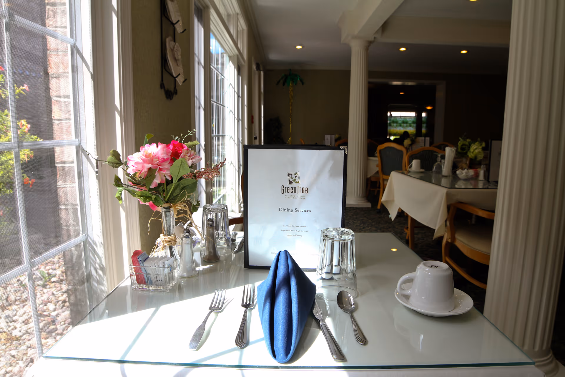 A dining table set for one with a blue folded napkin, silverware, an upside-down glass, a cup and saucer, and a menu labeled GreenTree Dining Services. A vase with pink flowers and condiments are also on the table. The room has large windows letting in natural light and other tables with chairs are visible in the background.