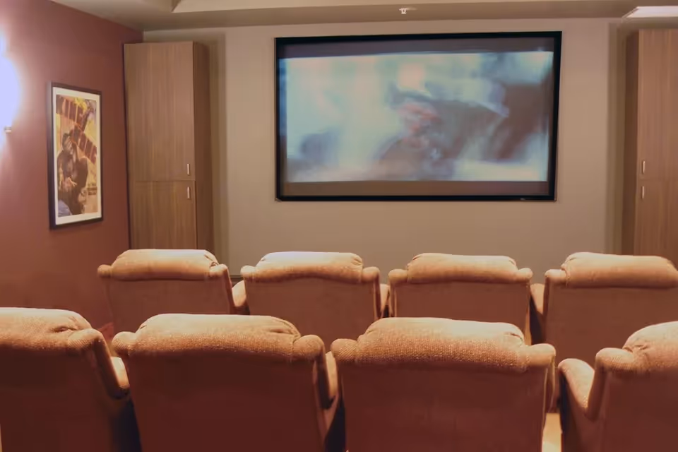 A small movie theater room with two rows of cushioned recliner chairs facing a large screen. The walls are painted in warm tones, with a framed King Kong movie poster on the left wall and wooden cabinets on either side of the screen.