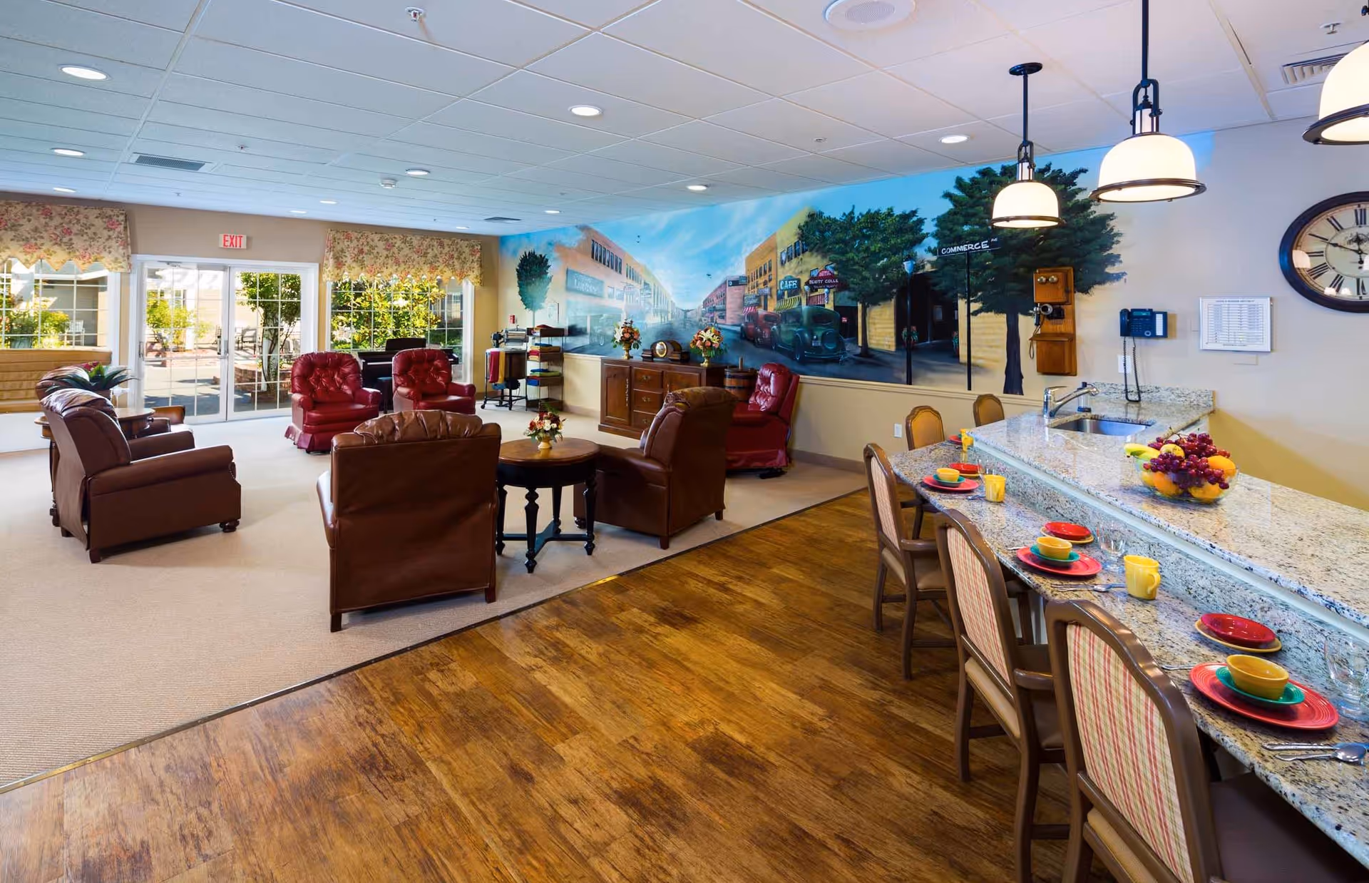 A cozy common area in a senior living facility featuring a seating area with brown and red leather armchairs arranged around small tables, a granite countertop with colorful place settings and chairs, and a large mural depicting a vintage street scene on the wall. The room has wood flooring near the counter and carpet near the seating area, with large windows and glass doors leading to an outdoor space.