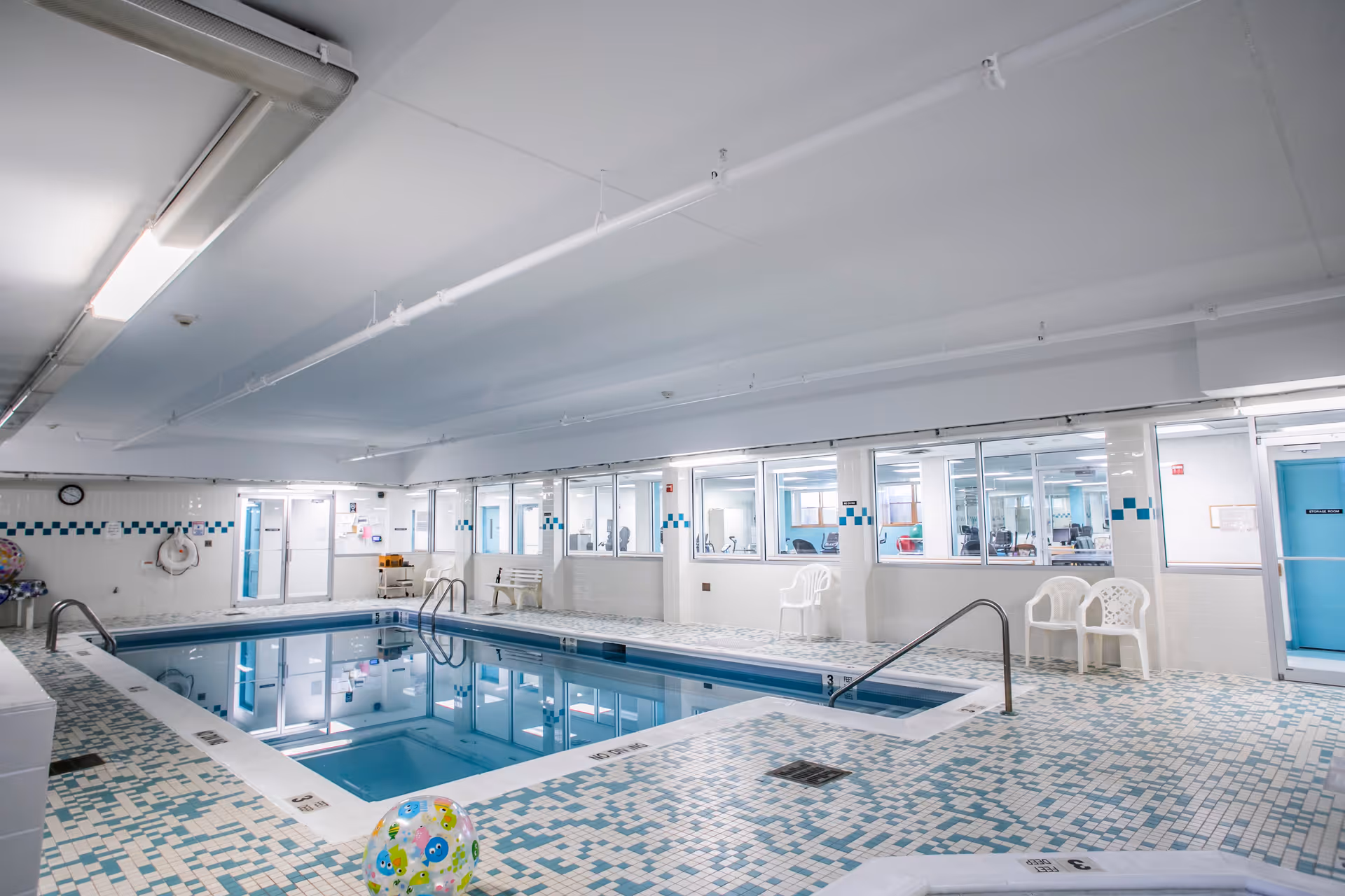 Indoor swimming pool area with tiled floor and walls, white plastic chairs along the side, and large windows looking into an adjacent room with exercise equipment.