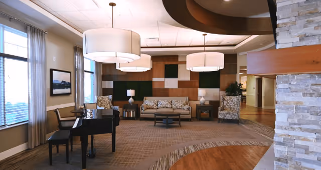 A spacious and well-lit living room area with large windows covered by sheer curtains, a black grand piano with a bench, patterned armchairs, a sofa with floral cushions, side tables with lamps, and modern circular ceiling lights. The room features a decorative wall with wooden panels and green accents, a stone column, and a mix of carpet and hardwood flooring.