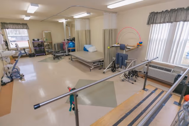 A rehabilitation therapy room with parallel bars for walking exercises, a treatment table with a pillow, exercise equipment including a stationary bike and therapy balls, and a window with vertical blinds and a valance.