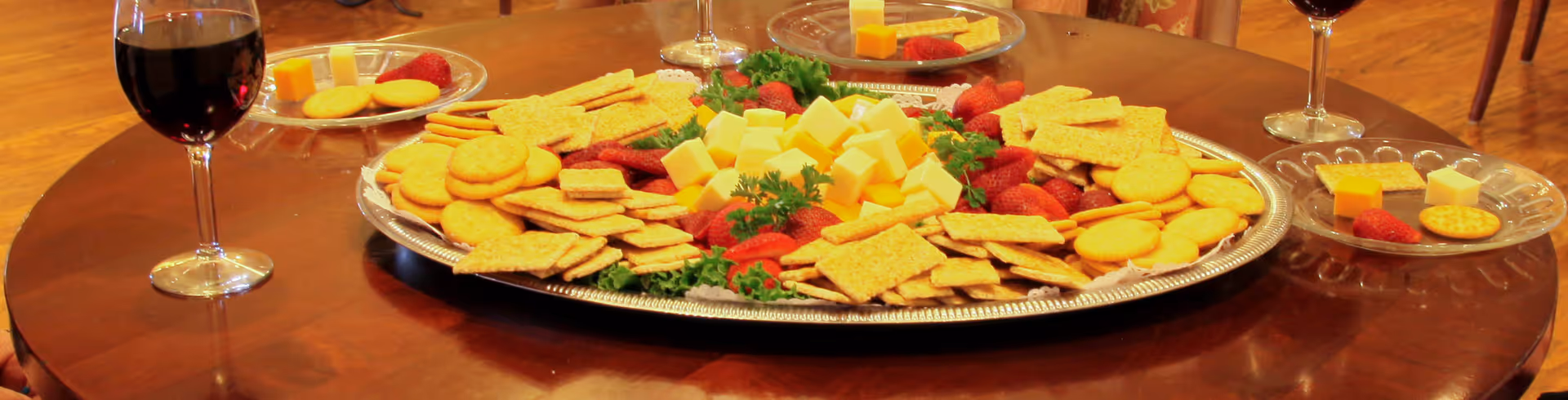 A wooden table set with a large silver platter filled with assorted crackers, cheese cubes, and strawberries. Surrounding the platter are smaller clear plates with similar snacks and glasses of red wine.