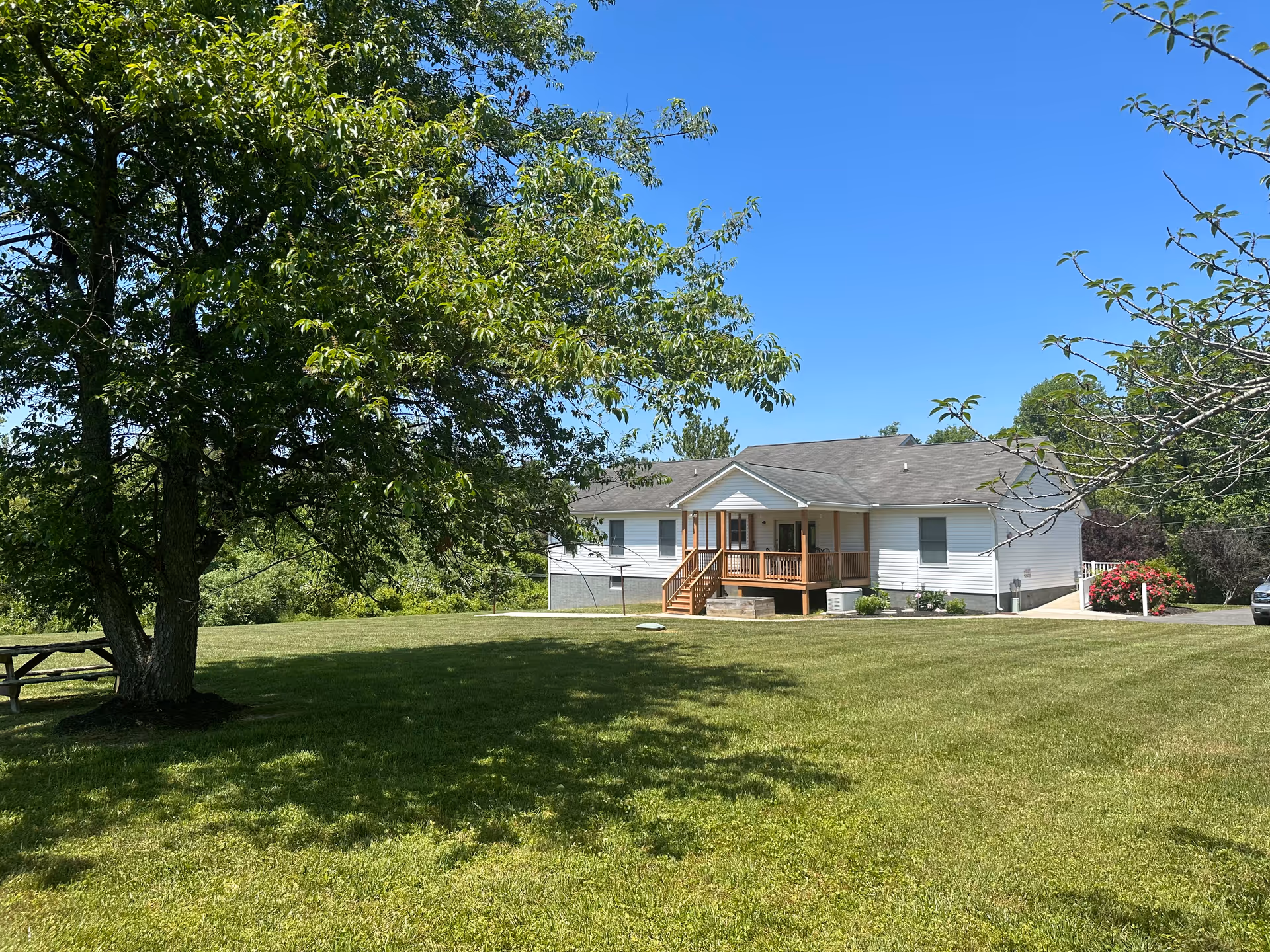 A single-story white building with a gray roof and a small wooden porch, surrounded by a large grassy lawn with trees and a picnic table under one tree, under a clear blue sky.