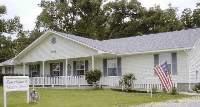 Single-story white building with a gray roof and a front porch with white railings. An American flag is displayed near the entrance. There is a sign in front of the building that reads 'Laurelwood Retirement Residence'. The building is surrounded by green grass and trees in the background.