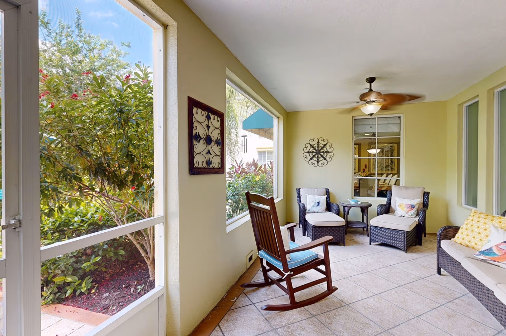 A cozy enclosed patio area with beige tiled floor, light yellow walls, and a ceiling fan. The space is furnished with a wooden rocking chair with a blue cushion, two wicker armchairs with matching ottomans and colorful pillows, a small round side table, and a wicker sofa with patterned cushions. Large windows and a screened door provide views of green bushes and trees outside.