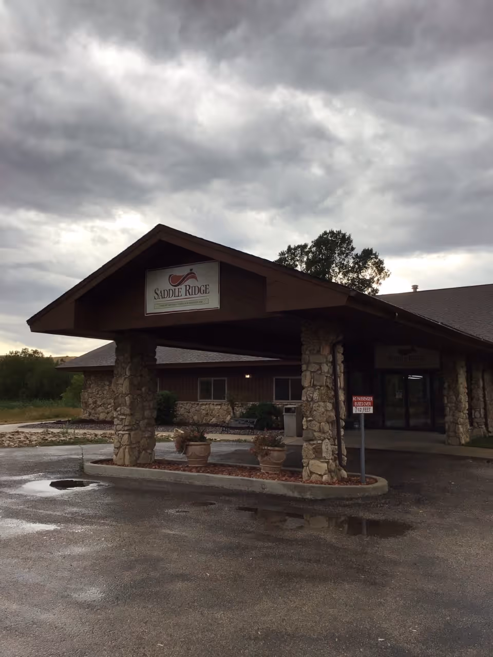 Covered stone entrance and porte-cochere of a senior living facility with a 'Saddle Ridge' sign under a cloudy sky.