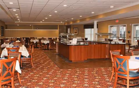 A spacious dining hall in a senior living facility with tables, chairs, and a service counter.
