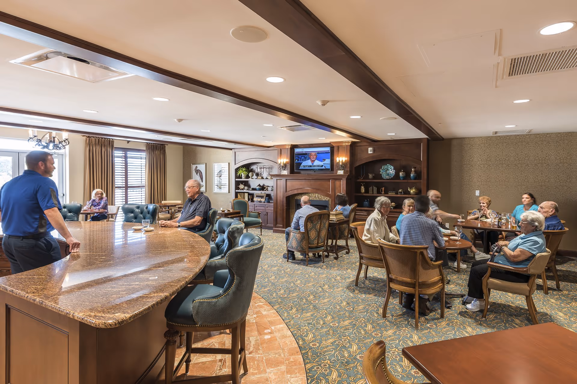 A spacious lounge area with a large bar, comfortable seating, and several groups of people engaging in conversation. A television is mounted on the wall, and decorative items are displayed on shelves.